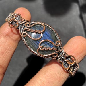 May include: A wire-wrapped bracelet featuring a central oval-shaped dark blue labradorite gemstone with iridescent streaks. The setting is crafted from oxidized copper wire, intricately woven into decorative swirls and loops along the band.