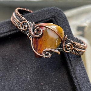 May include: A wire-wrapped ring featuring a polished, teardrop-shaped stone in shades of mustard yellow, brown, and deep red. The band is constructed from dark, oxidised copper wire, intricately coiled and woven into decorative spiral patterns.