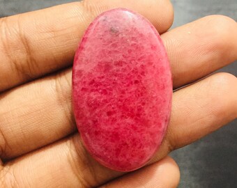 May include: An oval-shaped cabochon gemstone with a smooth, polished surface. The stone features a vibrant, deep pink color with a mottled, crystalline texture throughout, displaying natural variations in tone and small dark inclusions.