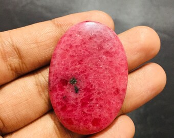 May include: An oval-shaped cabochon gemstone with a vibrant pink and magenta color palette. The stone features a textured, crystalline surface with small, dark inclusions scattered across its face, showcasing a polished and smooth finish.