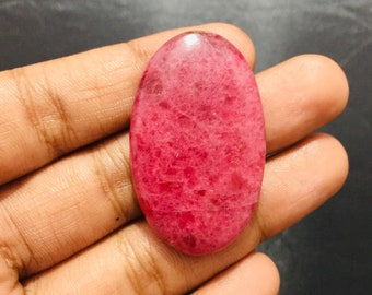 May include: An oval-shaped cabochon gemstone featuring a vibrant magenta and deep pink color palette with a natural mottled texture and a smooth, polished surface that reflects light across its rounded, symmetrical form.