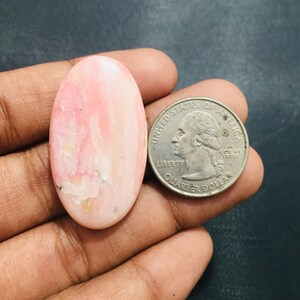 Pode incluir: Uma pedra preciosa de opala rosa polida, em forma oval, com veios brancos e rosa claro. Uma moeda de um quarto de dólar dos EUA está ao lado da pedra preciosa para comparação de tamanho.