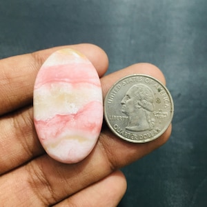 Pode incluir: Um cabochão de pedra preciosa oval com faixas rosa e creme, posicionado ao lado de uma moeda de 25 centavos de dólar americano para escala. A pedra apresenta camadas horizontais de rosa pastel suave e branco opaco, com acabamento polido.