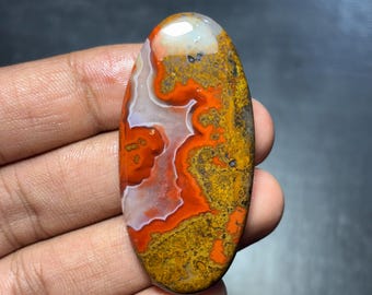 May include: An oval-shaped gemstone with a polished surface. The stone features a striking pattern of orange, white, and yellow-brown hues. The colors blend and swirl, creating a unique and natural design. The gemstone is held in a hand.