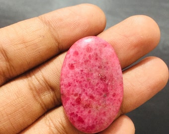 May include: An oval-shaped, polished gemstone with a vibrant pink and magenta mottled surface. The stone features a smooth, rounded top and a speckled texture with varying shades of deep red and light pink throughout its crystalline structure.