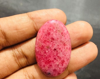 May include: An oval-shaped cabochon gemstone featuring a vibrant magenta and deep pink mottled surface with a polished, smooth finish. The stone displays a natural crystalline texture with small dark inclusions scattered throughout its surface.