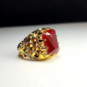 Pode incluir: Um anel de ouro com uma grande gema vermelha engastada em uma garra. A banda do anel é intrincadamente desenhada com um padrão de escamas.
