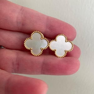 May include: A pair of gold-toned stud earrings, each featuring a four-leaf clover shape with a white mother-of-pearl inlay. The edges of the clover motifs are detailed with a beaded texture, creating a distinct border around the central white stone.