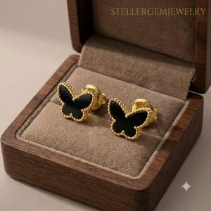 May include: A pair of gold stud earrings shaped like butterflies with black inlay and beaded edges. The earrings are displayed inside a dark brown wooden box with a tan velvet interior. The text STELLERGEMJEWELRY is visible at the top.