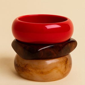 Peut inclure: Une pile de trois bracelets joncs. Le bracelet supérieur est d'un rouge vif uni et lisse. Le bracelet central est marron foncé avec une forme angulaire à facettes. Le bracelet inférieur est beige clair avec un motif marbré tourbillonnant.