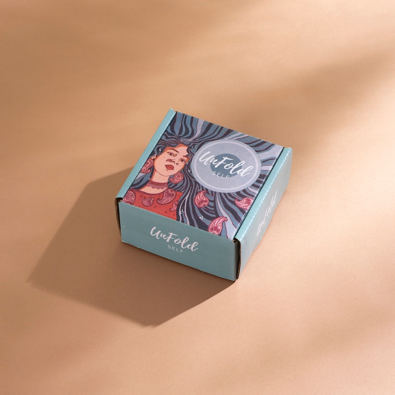 Puede incluir: Una peque&ntilde;a caja cuadrada con exterior turquesa y una colorida ilustraci&oacute;n en la parte superior. La ilustraci&oacute;n presenta a una mujer con cabello largo y suelto y las palabras "Unfold Self" en un logotipo circular y en la caja. La caja est&aacute; sobre una superficie beige.