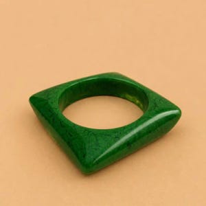 Pode incluir: Uma pulseira verde quadrada. A pulseira tem um acabamento brilhante e uma textura marmorizada. A joia está sobre um fundo neutro. A pulseira é um acessório arrojado.