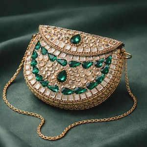 Puede incluir: Un bolso de mano dorado adornado con una cadena. El bolso está incrustado con piedras preciosas transparentes y verde esmeralda. Las piedras están dispuestas en un patrón decorativo en la parte delantera. El fondo es de tela verde oscuro.