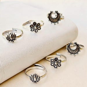 May include: A collection of silver-toned toe rings with various designs, including floral, crescent moon, and lotus flower motifs. The rings are displayed on a white surface, showcasing their intricate details and adjustable bands.