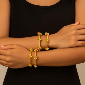 Könnte beinhalten: Vier goldfarbene Armreifen werden an Handgelenken gezeigt. Jeder Armreif hat ein gedrehtes Seildesign mit drei Clustern aus kleinen, runden Perlen. Der Schmuck ist vor einem dunklen Hintergrund platziert.