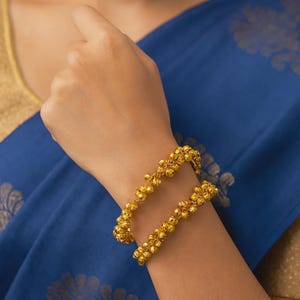 May include: A pair of gold-colored bangles, each adorned with small, spherical embellishments. The bangles are worn on a wrist, set against a backdrop of blue fabric. The jewelry appears to be made of metal and reflects light.