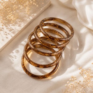 Puede incluir: Un conjunto de cinco pulseras marrones translúcidas con un diseño veteado. Las pulseras brillantes están apiladas sobre una superficie crema. Flores secas y una tela blanca doblada en el fondo. Cada pulsera tiene un diámetro de aproximadamente 6 cm.