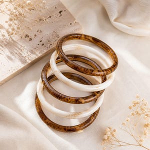 Puede incluir: Una pila de cinco pulseras redondas, alternando marrón y crema. Las pulseras marrones presentan un efecto veteado, mientras que las otras son de color crema liso. Las joyas se presentan sobre una tela blanca, con un bloque de piedra en el fondo.