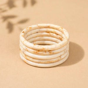 May include: A stack of six ivory-coloured bangles with brown marbling. The bracelets are round and smooth, stacked on top of each other. The background is a solid tan colour.