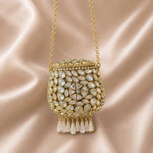 Pode incluir: Uma pequena bolsa arredondada em tom dourado com um exterior intrincado coberto por cristais transparentes em forma de gota e um motivo floral central. A bolsa inclui uma alça de corrente dourada longa e pingentes de cristal na base.