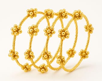 May include: Four gold-colored bangles with a floral design. Each bangle features a twisted rope-like band and is adorned with clusters of small, round beads arranged in a flower pattern. The jewelry is set against a white background.