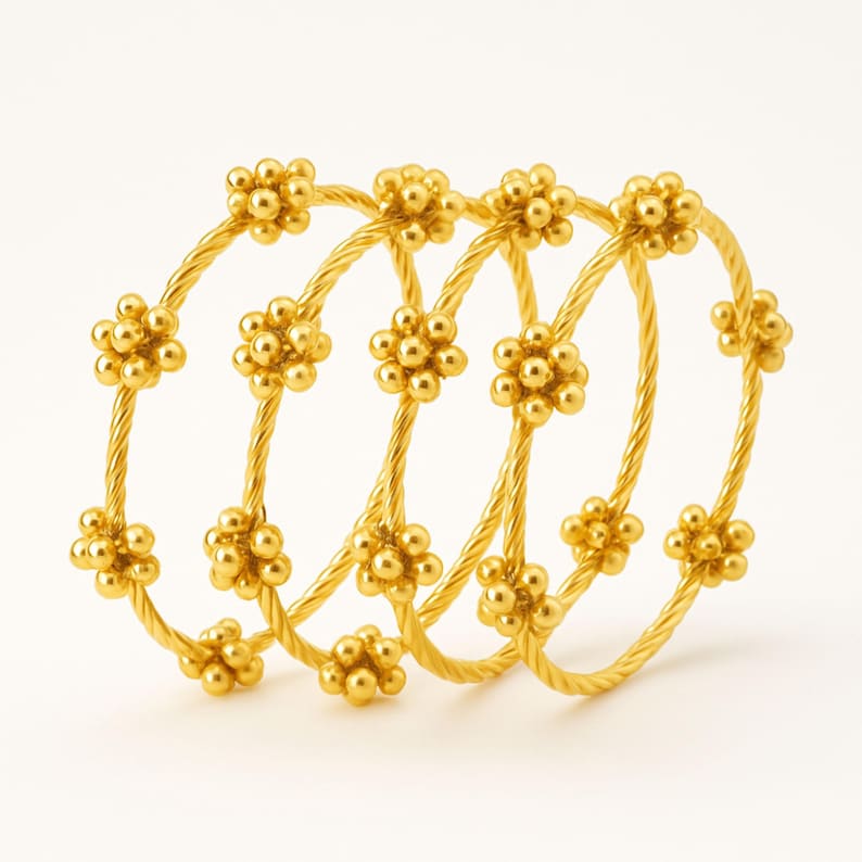 Puede incluir: Cuatro pulseras doradas con un dise&ntilde;o floral. Cada pulsera presenta una banda trenzada en forma de cuerda y est&aacute; adornada con racimos de peque&ntilde;as cuentas redondas dispuestas en forma de flor. Las joyas est&aacute;n sobre un fondo blanco.