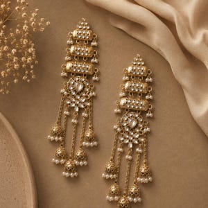 May include: A pair of gold-toned chandelier earrings. Each earring features multiple tiers of clear, sparkling stones and small, white pearls. Dangling chains lead to bell-shaped charms with more pearls, creating an elegant, ornate design.
