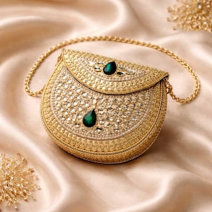Pode incluir: Uma clutch dourada semicircular com uma corrente. A bolsa é ricamente adornada com pedras claras e brilhantes e duas pedras preciosas verdes esmeralda em forma de gota. O fundo é um tecido macio e acetinado.