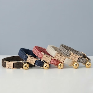 Op de afbeelding: Vier huisdierhalsbanden in bruin, blauw, rood en crème met een visgraatpatroon. Elke halsband heeft een goudkleurige metalen gesp gegraveerd met een naam en telefoonnummer, samen met een kleine, ronde goudkleurige bel aan de zijkant.