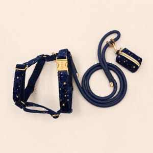 May include: A navy blue pet harness and matching rope lead featuring a gold star pattern. The harness includes a gold buckle with the text COCO, and the set comes with a small rectangular pouch in the same star-patterned fabric with gold hardware.
