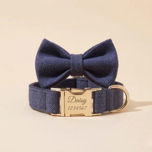 May include: A navy blue dog collar and bow tie set. The collar has a gold-coloured metal buckle engraved with the name "Daisy" and the number "1234567". The bow tie is attached to the collar. The set is made of a textured fabric.