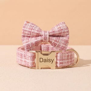 May include: A pink and white plaid pet collar featuring a matching bow tie and a gold-toned metal buckle engraved with the name Daisy. The collar includes a gold-toned metal D-ring for attaching a lead, showcasing a textured fabric construction.