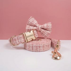 Op de afbeelding: Een roze en witte tweed halsband voor huisdieren met een bijpassende strik en riem. De halsband is voorzien van een goudkleurige metalen gesp met de tekst SWING, een gouden D-ring en een gouden musketonhaak aan de opgerolde riem.