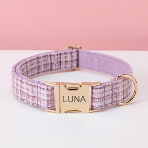 May include: A pet collar featuring a purple and white plaid fabric pattern with a gold-toned metal side-release buckle. The buckle is engraved with the name LUNA in a dark font. The collar includes a gold-toned metal D-ring for attaching a lead.