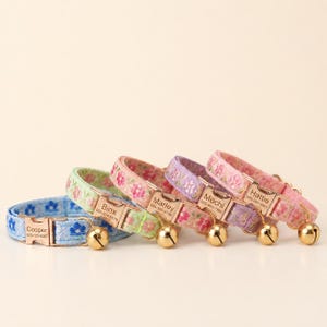 May include: Four pet collars in blue, green, pink, and purple with floral patterns. Each collar features a rose gold metal buckle engraved with a name and phone number, including Cooper, Binx, Marley, Mochi, and Hattie, plus a small bell.