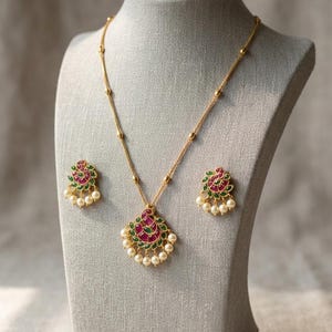 May include: A gold necklace and earring set on a display bust. The pendant and earrings have a fan-shaped design with red and green detailing, and small white pearls. The necklace chain features small gold beads.
