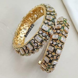 Peut inclure: Deux bracelets dorés ornés de pierres claires rectangulaires et rondes et d'accents verts. Le mot "BANGLES" est imprimé sur le côté d'un bracelet. Les bijoux sont présentés sur fond blanc.