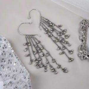 Puede incluir: Un par de puños de oreja plateados con cadenas colgantes y pequeñas cuentas redondas. Los puños de oreja tienen un diseño curvo que se engancha sobre la oreja. Las cadenas caen en cascada, creando un efecto en capas.