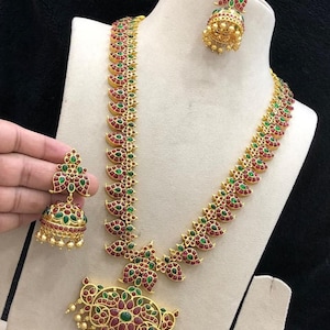 May include: Antique gold-colored brass metal jewelry set with red and green Kempu stones. The set includes a long necklace, a pair of earrings, and a pendant. The necklace and earrings have a detailed design. The text on the image says "PC 1545".