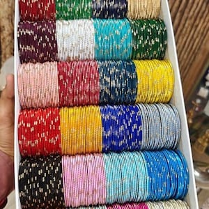 May include: A box filled with multiple rows of colorful bangles. The bangles are in various colors, including red, green, blue, gold, and pink. Each bangle has a metallic sheen and is arranged neatly in the box, creating a vibrant display.