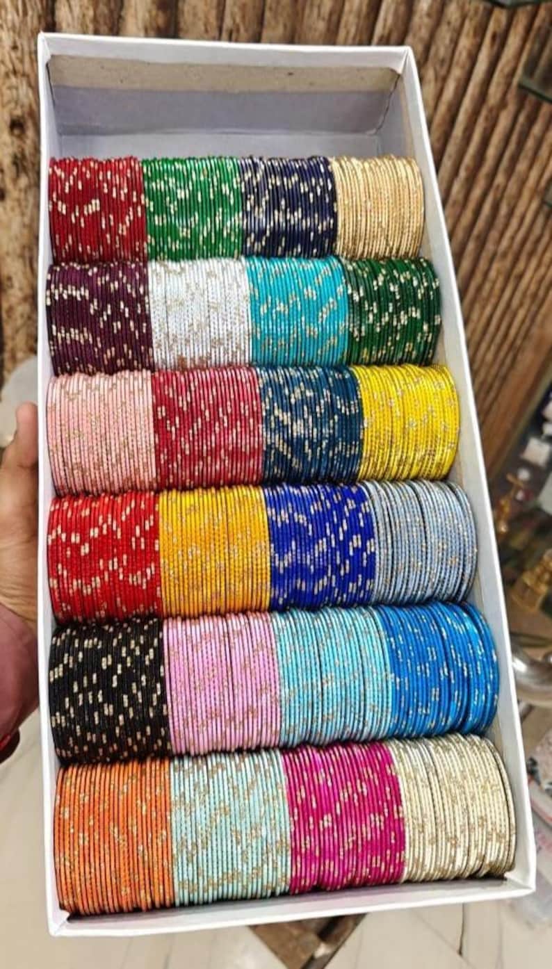 May include: A box filled with multiple rows of colorful bangles. The bangles are in various colors, including red, green, blue, gold, and pink. Each bangle has a metallic sheen and is arranged neatly in the box, creating a vibrant display.