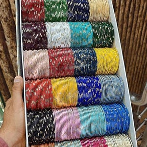May include: A box filled with stacks of colorful bangles. The bangles are in various colors, including red, green, blue, yellow, and pink. Each bangle is adorned with a subtle, shimmering pattern. The box is held by a person.