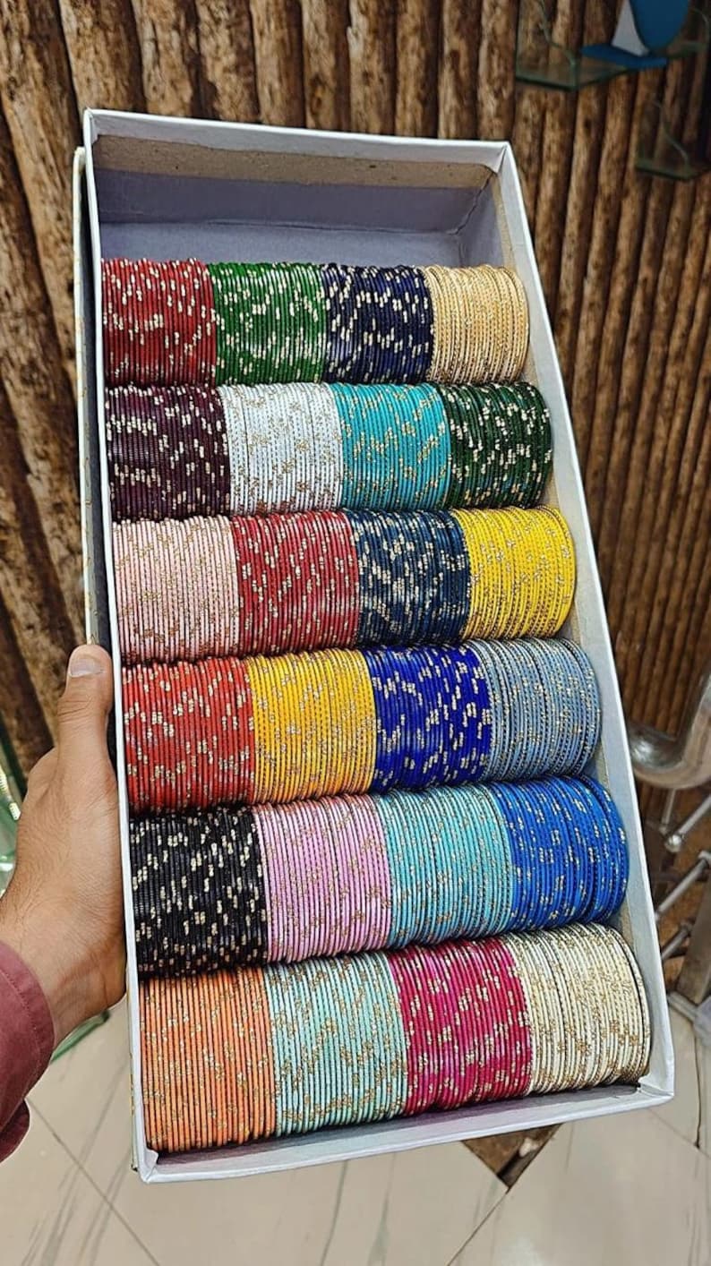 May include: A box filled with stacks of colorful bangles. The bangles are in various colors, including red, green, blue, yellow, and pink. Each bangle is adorned with a subtle, shimmering pattern. The box is held by a person.