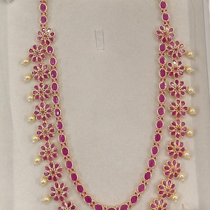 Peut inclure: Un ensemble collier et boucles d'oreilles dorées. Le collier présente un design à double rangée avec des pierres de couleur rubis ovales et des motifs floraux. De petites perles pendent des éléments floraux. Les boucles d'oreilles reprennent le design du collier.