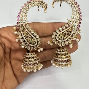 May include: A pair of gold-tone ear cuffs featuring a large paisley-shaped frame encrusted with clear stones, pink accents, and small green details. Each piece includes a dangling bell-shaped jhumka pendant adorned with pearls and stones.