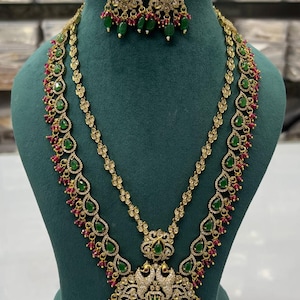 May include: A gold-toned jewelry set featuring a necklace and matching earrings. The necklace has a layered design with green and red accents, and a pendant with a detailed peacock design. The earrings have a similar design with green and red accents.