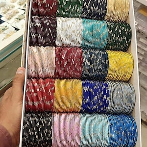 May include: A box filled with multiple stacks of colorful bangles. The bangles are in various colors, including red, green, blue, yellow, and pink, with gold accents. The bangles are stacked neatly in rows, creating a vibrant display of jewelry.