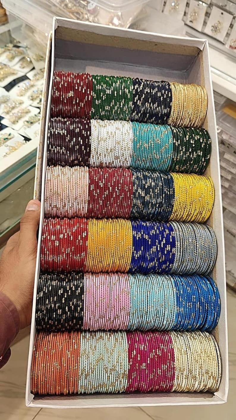 May include: A box filled with multiple stacks of colorful bangles. The bangles are in various colors, including red, green, blue, yellow, and pink, with gold accents. The bangles are stacked neatly in rows, creating a vibrant display of jewelry.