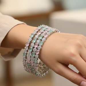 Puede incluir: Una pulsera plateada con filas alternas de piedras preciosas ovaladas rosas y verdes. La pulsera está adornada con pequeñas piedras de acento transparentes, creando un efecto brillante. La joya se lleva en la muñeca.