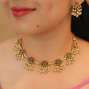 May include: Gold-toned necklace and earring set. The jewelry features floral designs with red and green accents, and small white pearl-like beads. The necklace and earrings are worn on a person.